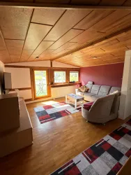 Ein modernes Wohnzimmer mit Holzdecke und großen Fenstern. Es ist gemütlich eingerichtet mit einem grauen Sofa und einem Tisch.