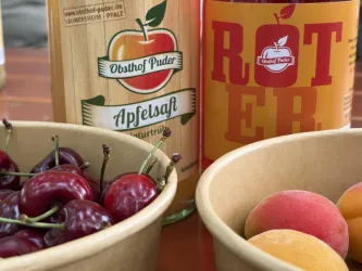 Ein Tisch mit frischen Kirschen und Aprikosen in Schalen. Neben den Früchten stehen Flaschen mit Apfelsaft und einem Getränk namens "Roter".