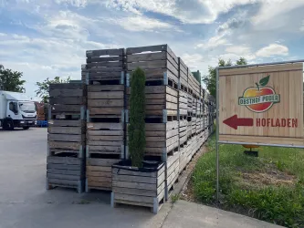 Lager von Holzpaletten neben einem Schild mit der Aufschrift "Hofladen". Im Hintergrund ist ein Lkw zu sehen und der Himmel ist teilweise bewölkt.