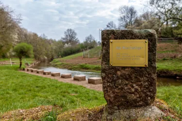 Ein ruhiger Flusslauf mit Steinen im Wasser und grünem Gras. Im Vordergrund steht ein Stein mit einer Plakette.
