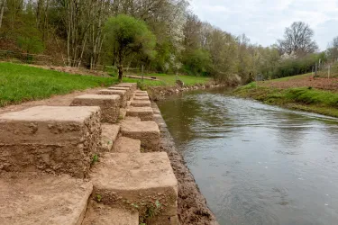 Ein ruhiger Fluss mit angrenzenden, grünen Wiesen. Am Ufer sind große Steine zu sehen, die als Stufen ausgelegt sind.
