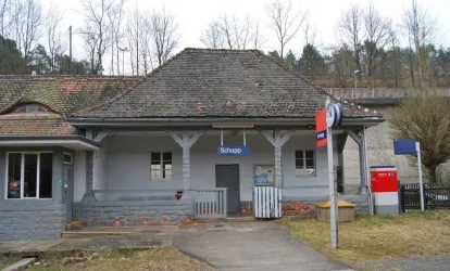 Ein älterer Bahnhof in Schoop mit einem schlichten, grauen Gebäude. Im Vordergrund sind ein Parkplatz und einige Bäume zu sehen.