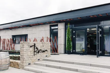 Ein modernes Gebäude mit einem stilvollen Eingangsbereich und der Beschriftung "Bram". Vor dem Eingang gibt es eine Treppe und dekorative Elemente aus Stein und Pflanzen.