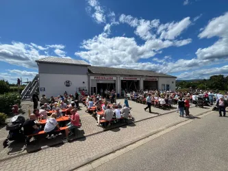 Eine große Menschenmenge sitzt an den Tischen vor einem Gebäude. Das Wetter ist sonnig mit vielen Wolken am Himmel.