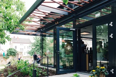 Ein modernes Café mit großen Fenstern und einem einladenden Außenbereich. Grünpflanzen und Blumen sorgen für eine angenehme Atmosphäre.