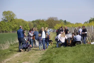 Eine Gruppe von Menschen versammelt sich auf einem Feldweg, um gemeinsam Zeit zu verbringen. Einige sitzen an Tischen, während andere stehen und sich unterhalten.