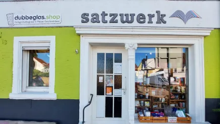 Ein Geschäft mit einer bunten Fassade in grüner Farbe. Der Name "satzwerk" ist über dem Eingang zu sehen und das Schaufenster zeigt verschiedene Bücher und Produkte.