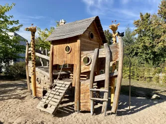 Ein kreatives Spielhaus aus Holz mit einer Rutsche und Kletterelementen. Es hat zwei kleine Giraffenfiguren an den Seiten und ist von Bäumen umgeben.
