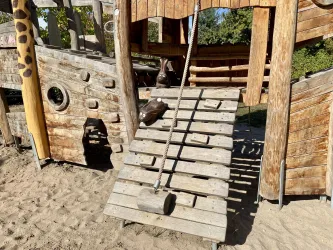 Ein Spielplatz mit einer Holzstruktur und einer erhöhten Plattform. Eine Schaukel und eine Rampe sind gut sichtbar.