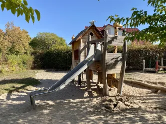 Ein Spielplatz mit einer Rutsche und einem kleinen Spielhaus. Der Bereich ist von Bäumen umgeben und hat einen Sandboden.