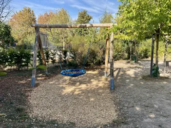 Eine Schaukel aus Holz steht in einem Spielplatzbereich mit Kieselboden. Umgeben von Bäumen und Sträuchern bietet der Platz eine ruhige Atmosphäre.