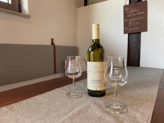 Eine Flasche Weißwein steht auf einem Tisch mit zwei Weingläsern. Im Hintergrund ist eine Wand mit einem handschriftlichen Schild zu sehen.