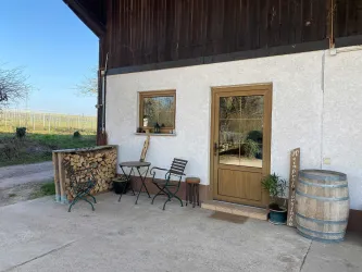 Ein gemütlicher Außenbereich eines Hauses mit einem Eingang und einem Fenster. Daneben steht ein Tisch mit Stühlen und ein Holzstapel.