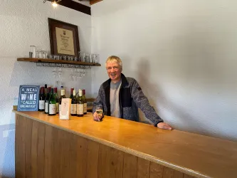 Ein freundlicher Mann steht an einer Holzbar und hält ein Glas Getränk in der Hand. Hinter ihm sind verschiedene Weinflaschen und Gläser auf einem Regal angeordnet.