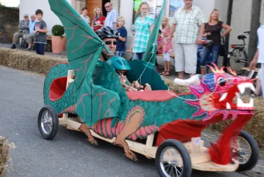 Ein kreatives Fahrzeug in Form eines Drachen, das von einem Erwachsenen und einem Kind gefahren wird. Im Hintergrund stehen Zuschauer, die das Rennen beobachten.