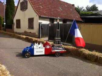 Ein kleines, dekoriertes Auto in den Farben der französischen Flagge steht an einer Straße. Im Hintergrund sind ein paar Häuser und eine Fahne zu sehen.