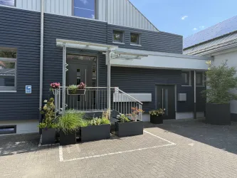 Ein modernes Gebäude mit einem Eingangsbereich und einer überdachten Treppe. Vor dem Gebäude befinden sich große Pflanzkübel mit bunten Blumen und grünen Pflanzen.