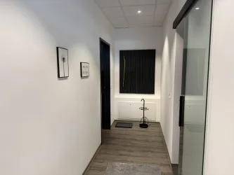 Ein moderner Flur mit weißer Wandfarbe und Holzfußboden. Rechts sind Glastüren und links hängen Bilder an der Wand.