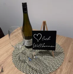 Ein Willkommensgruß mit einem Flasche Wein, zwei Gläsern und einem kleinen Schild. Die Atmosphäre ist gemütlich und einladend.
