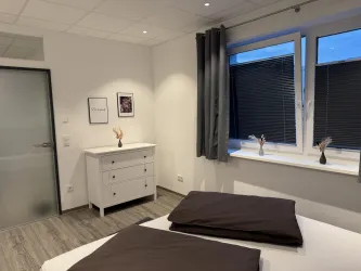 Ein modernes Schlafzimmer mit einem Doppelbett und zwei Nachttischen. Große Fenster lassen viel Tageslicht herein und es gibt dekorative Elemente auf der Kommode.