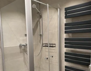 Eine moderne Dusche mit Glaswänden und einer Handbrause. An der Wand befindet sich ein Heizkörper in dunkler Farbe.