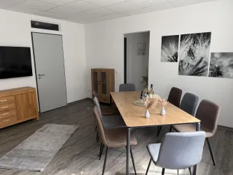 Ein modernes Esszimmer mit einem großen Tisch und sechs Stühlen. An der Wand hängen Kunstwerke, und eine TV-Einheit ist sichtbar.