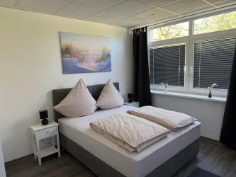 Ein modernes Schlafzimmer mit einem großen Bett und zwei dekorativen Kissen. An der Wand hängt ein Bild eines Winterlandschafts, und es gibt große Fenster, die viel Tageslicht hereinzulassen.
