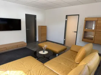 Ein modernes Wohnzimmer mit einem auffälligen, gelben Sofa und einem eleganten Couchtisch. Die Wände sind hell und es gibt eine große Wand mit einem Fernseher und Holzregalen.