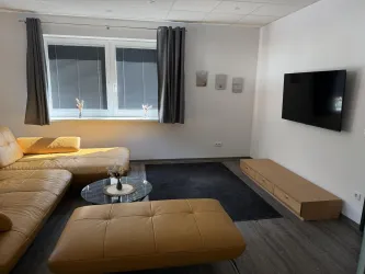 Ein modernes Wohnzimmer mit einem beigen Sofa und einem Glastisch. An der Wand hängt ein Fernseher, und es gibt große Fenster mit Vorhängen.