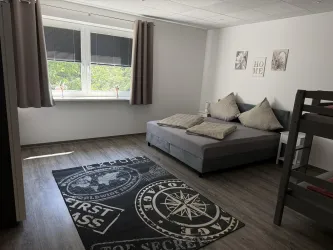 Ein helles Zimmer mit zwei Fenstern und einem großen Bett. Es gibt einen gemütlichen Teppich und Kissen auf dem Bett.
