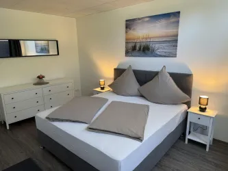Ein modernes Schlafzimmer mit einem Doppelbett und großen Kissen. An der Wand hängt ein Kunstwerk mit einem Strandmotiv, und es gibt Nachttische mit Lampen.
