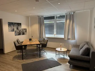 Ein modernes Wohnzimmer mit einem Esstisch und Stühlen. Es gibt ein graues Sofa und große Fenster mit Vorhängen.