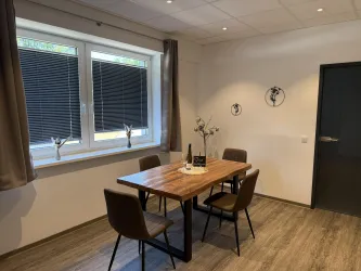 Ein moderner Essbereich mit einem Holz-Tisch und vier Stühlen. Die Fenster lassen viel Tageslicht herein und es gibt dezente Wanddekorationen.