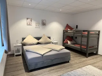 Ein modernes Schlafzimmer mit einem Doppelbett und einem Etagenbett. Die Wände sind in neutralen Farben gestaltet, und es gibt dekorative Bilder.