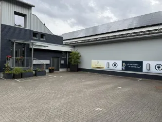 Ein modernes Gebäude mit einem gepflasterten Parkplatz. Im Hintergrund sind Werbetafeln und ein Solardach zu sehen.