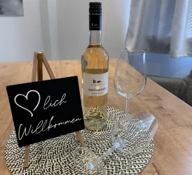 Ein Willkommensgeschenk mit einer Flasche Weißwein und Gläsern auf einem Tablett. Ein Schild mit der Aufschrift "Herzlich Willkommen" ergänzt die gemütliche Atmosphäre.