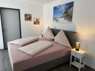 Ein gemütliches Schlafzimmer mit einem Doppelbett und großen Kissen. An der Wand hängen Bilder von einem Strand und der Raum ist hell und einladend.