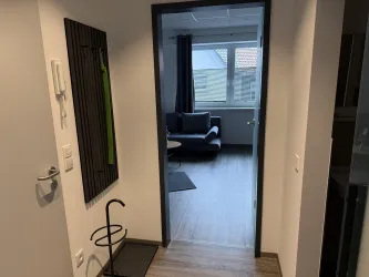 Ein Flur mit einem Blick auf ein Zimmer. Im Zimmer steht ein gemütliches Sofa und es gibt Tageslicht durch ein Fenster.
