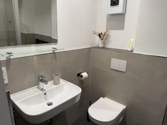 Ein modernes Badezimmer mit einem Waschbecken und einer Toilette. Die Wände sind in neutralen Farbtönen gehalten und es gibt einen kleinen Spiegel.