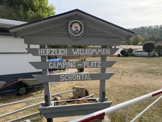 Ein ruhiger See umgeben von Bäumen und Wiese. Menschen genießen die Sonne und das Wasser.