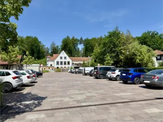 Ein Parkplatz mit mehreren Autos vor einem weißen Gebäude. Im Hintergrund sind Bäume und ein klarer blauer Himmel zu sehen.