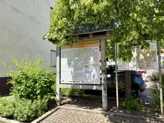 Eine Informationsstätte mit einer Karte steht im Freien, umgeben von Bäumen und Pflanzen. Im Hintergrund ist ein Auto zu sehen.