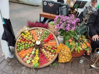 Ein dekorativer Obst- und Gemüsehaufen auf einem Wagenrad, umgeben von bunten Blumen. Die Frucht- und Gemüseanordnung zeigt eine Vielzahl von Farben und Formen.