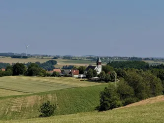 Eine malerische Landschaft mit sanften Hügeln und Feldern. Im Vordergrund steht eine Kirche, umgeben von kleinen Häusern und Bäumen.