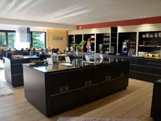 Ein modernes Küchenstudio mit großen, dunklen Kücheninseln und hellen Wänden. Im Hintergrund sind Regale mit Küchenutensilien und eine einladende Atmosphäre zu sehen.