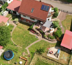 Ein großes Haus mit rotem Dach und gepflegtem Garten ist zu sehen. Der Garten umfasst eine Terrasse, einen Spielplatz und einen Trampolin.