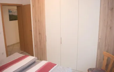 Ein einfach eingerichtetes Schlafzimmer mit zwei Betten und einem weißen Kleiderschrank. Im Hintergrund ist eine Holzoptik an der Wand zu sehen.