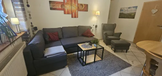 Ein gemütliches Wohnzimmer mit einer grauen Couch und einem Couchtisch. An der Wand hängen Bilder und es gibt eine Stehlampe sowie einen kleinen Esstisch.