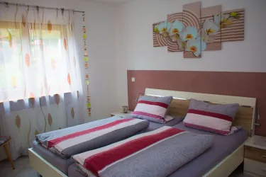 Ein gemütliches Schlafzimmer mit einem Doppelbett und bunten Kissen. Die Fenster sind mit hellen Vorhängen verhängt und es gibt ein dekoratives Wandbild.