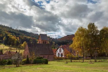 Eine malerische Landschaft mit einer kleinen Kirche und traditionell-bäuerlichen Häusern. Im Hintergrund sind sanfte Hügel und bunten Herbstbäume zu sehen.